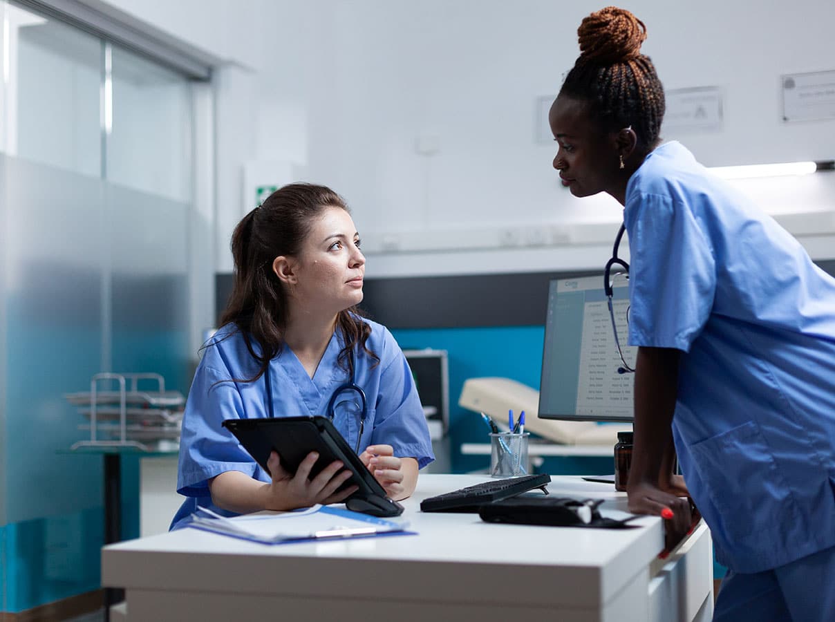 charge nurse standing over a desk at a clinic listening to another nurse give an excuse for nurses to call in sick