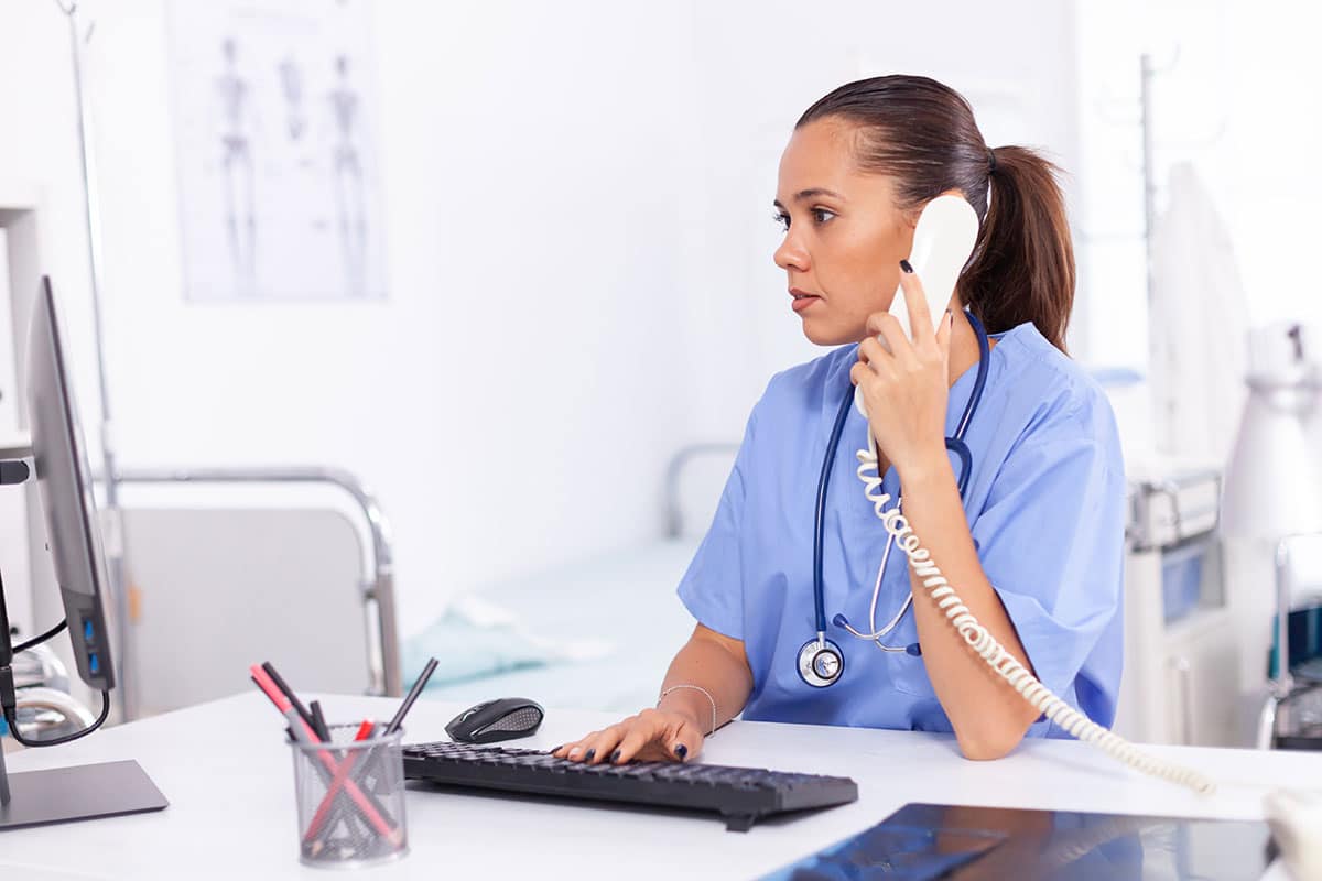 nurse in light blue robe sitting at a reception desk, on the phone taking a call from a coworker who is calling in sick