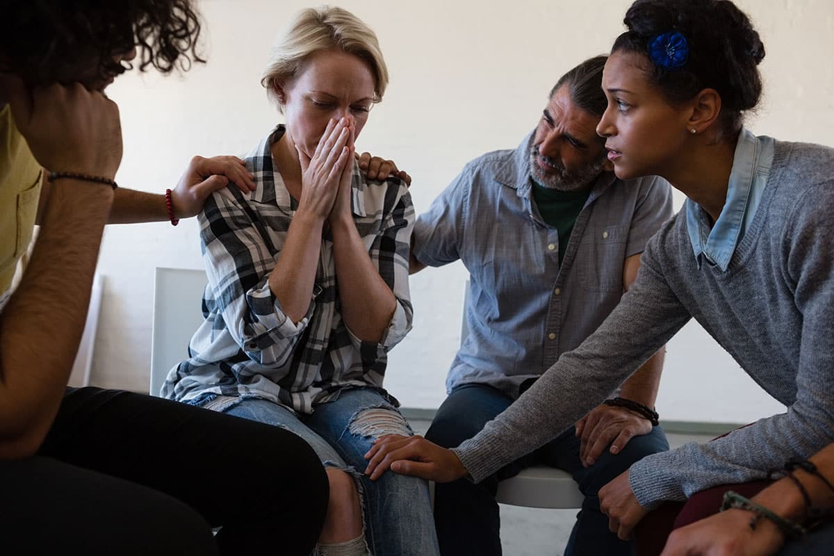 blonde woman has her hands to her mouth while wearing a plaid shirt as she is being consoled by a group of friends