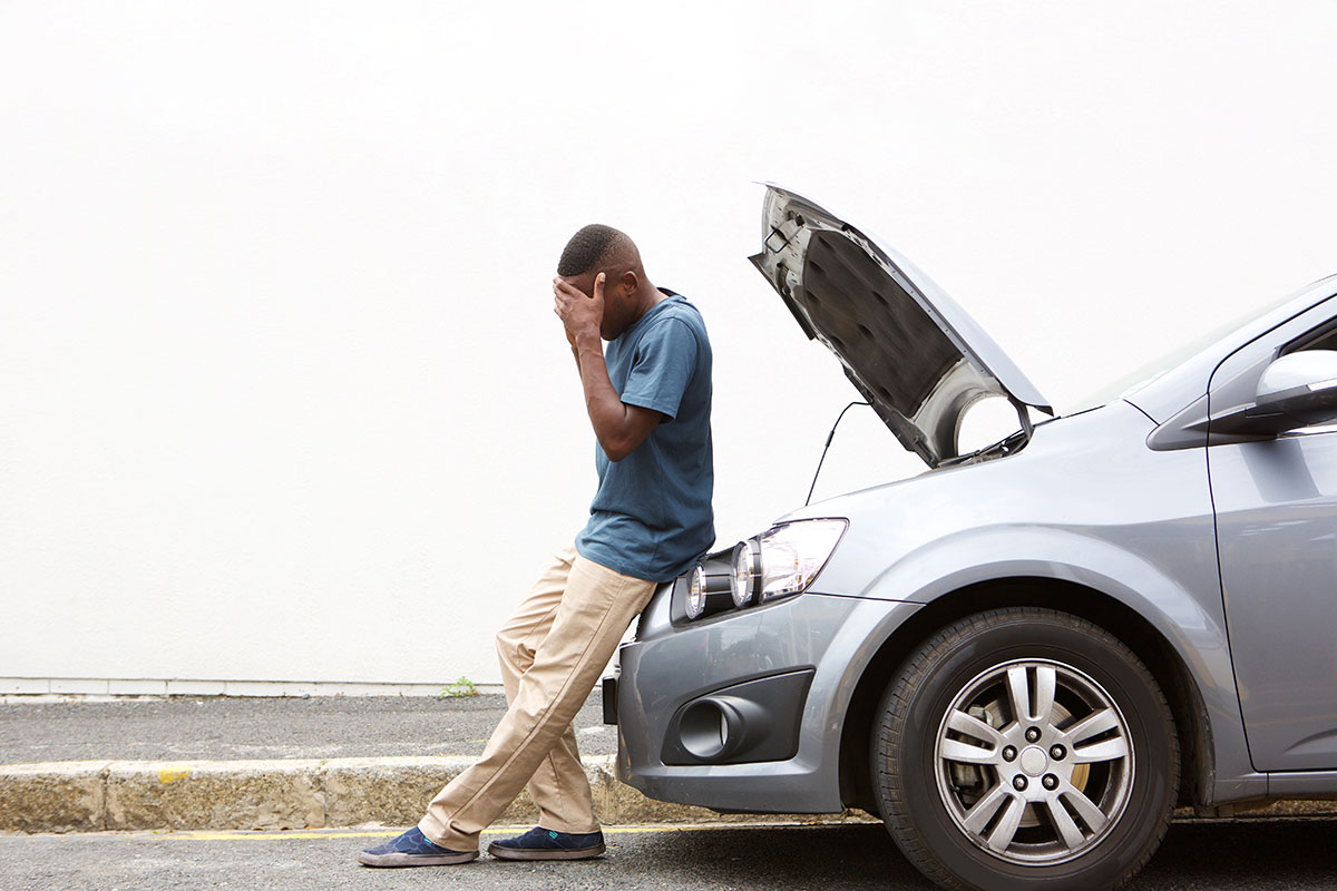 man with face in his hands sitting on the bumper of his car with the hood up because the car broke down