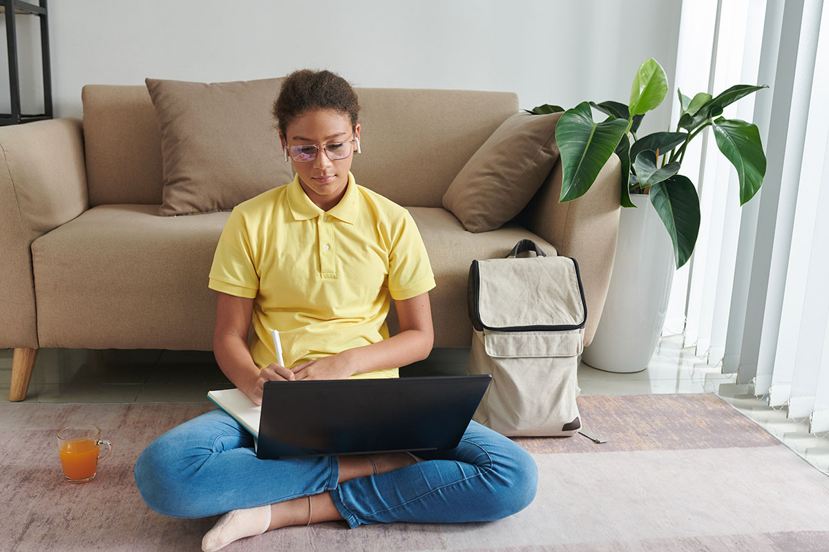 women in a yellow shirt and jeans is sitting cross-legged on the floor doing an online learning program while thinking of excuses for missing online school