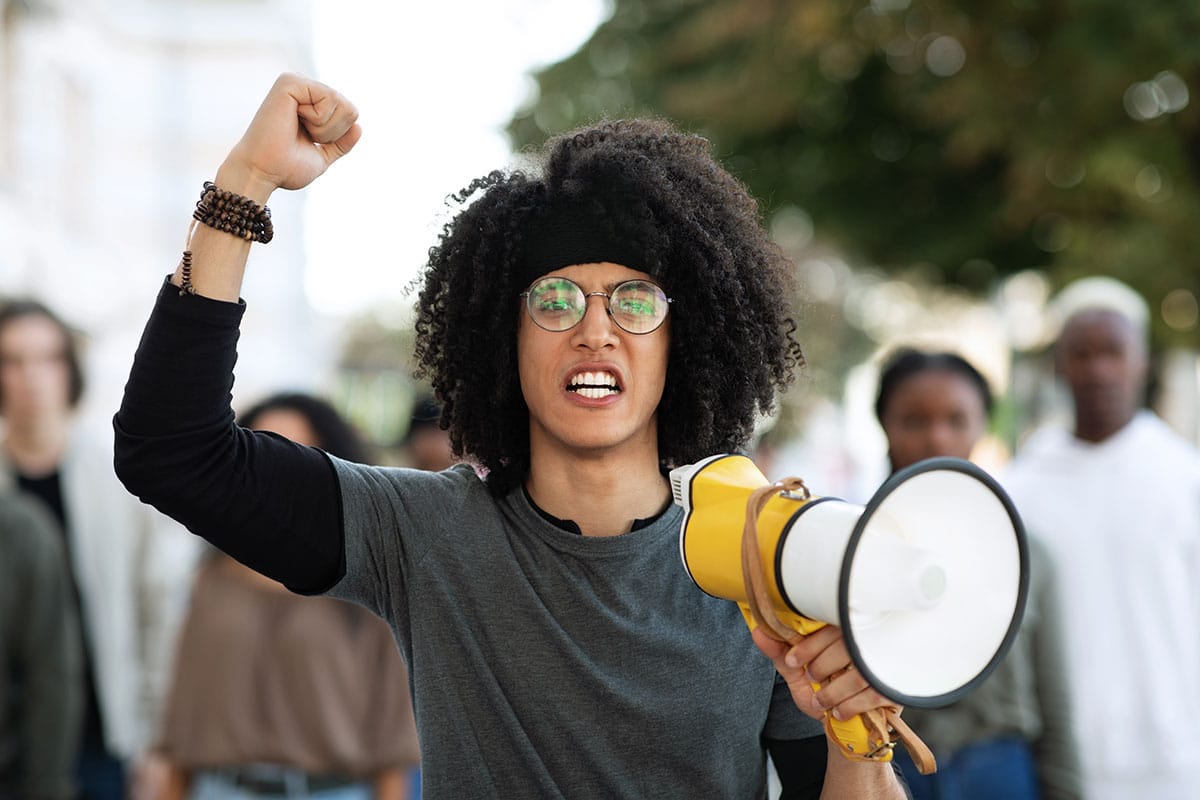 weirdo with an afro and round glasses has his fist in the air and a megaphone in his hand at a protest