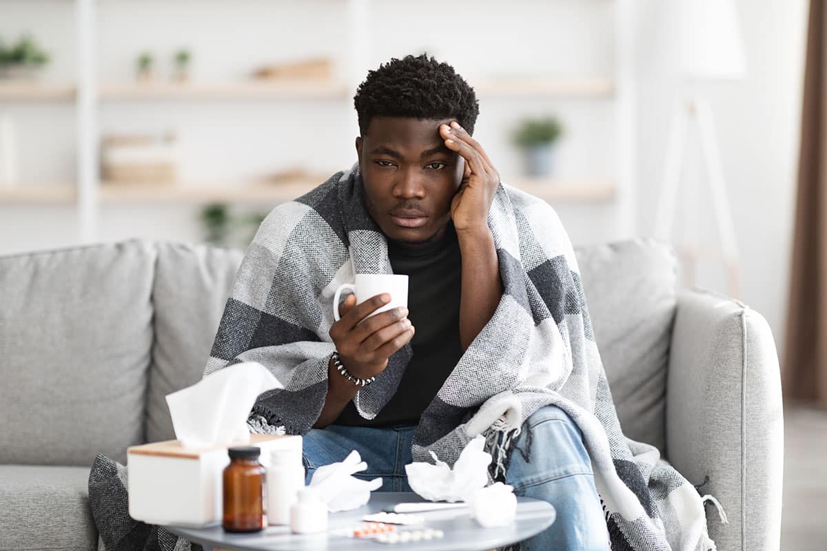 black man sits on a couch drinking tea while surrounded by kleenex and medications because he has the flu