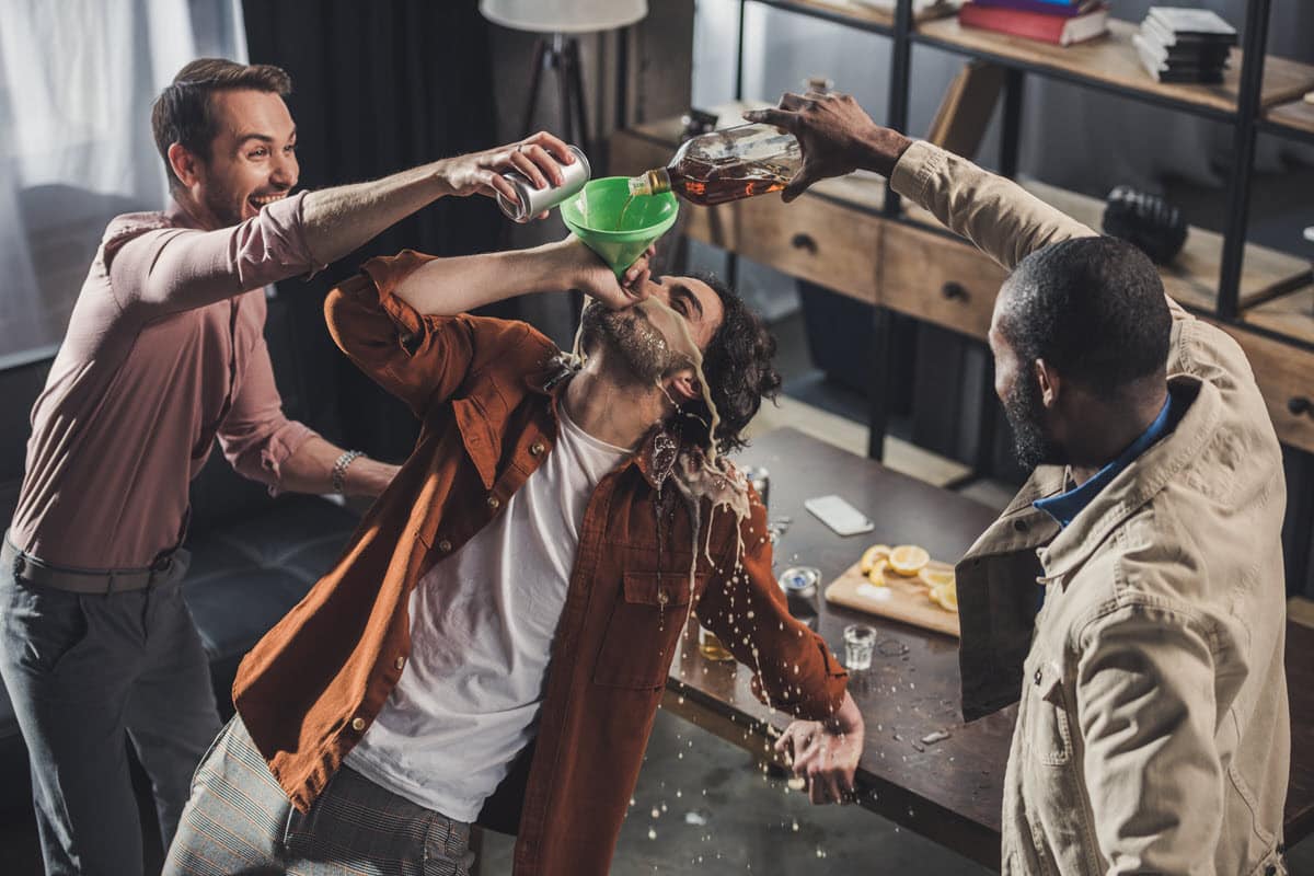 two guys pouring alcohol into a funnel that a third guy is drinking while spilling it all over himself