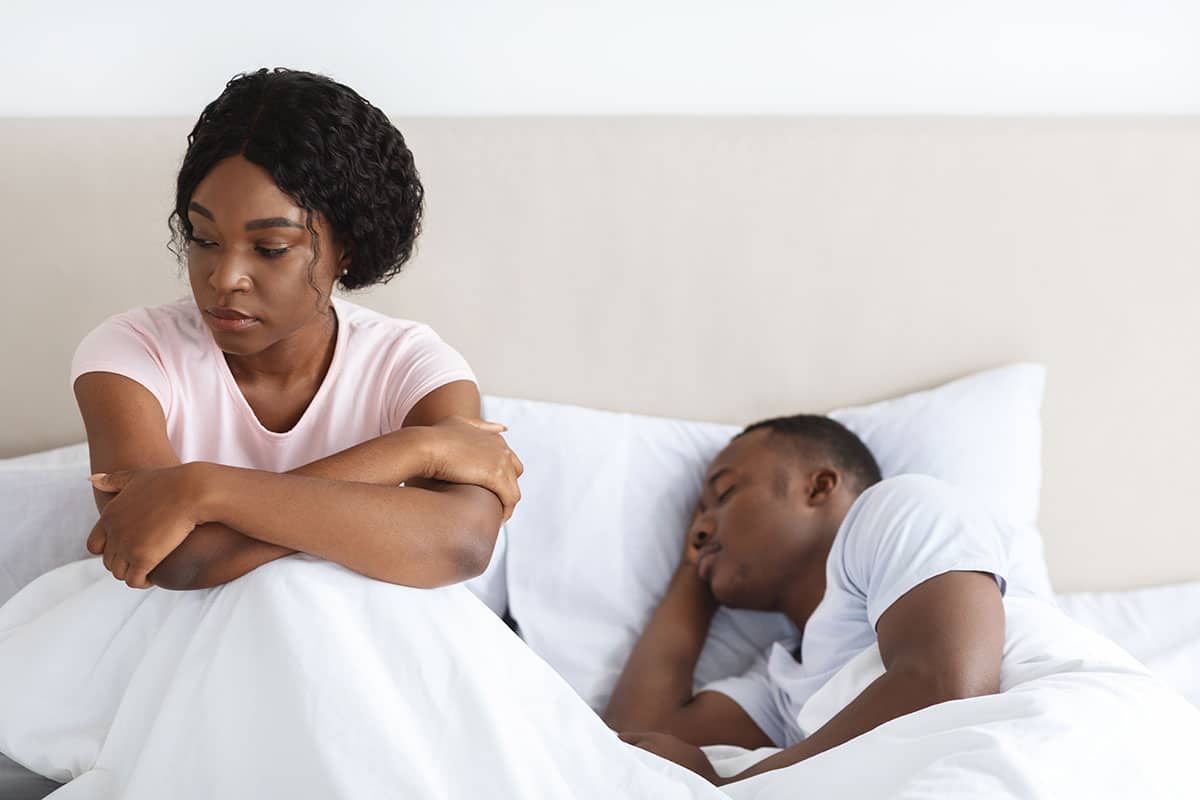 african woman sitting in bed upset because her husband fell asleep instead of throwing her the bone