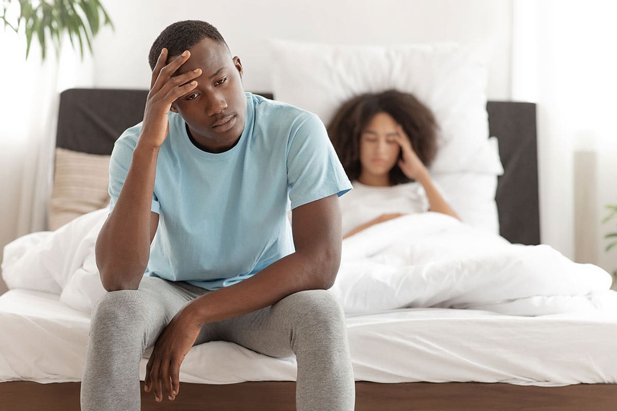 man sits at the foot of the bed frustrated because his girlfriend gave him the headache version of excuses to not have sex