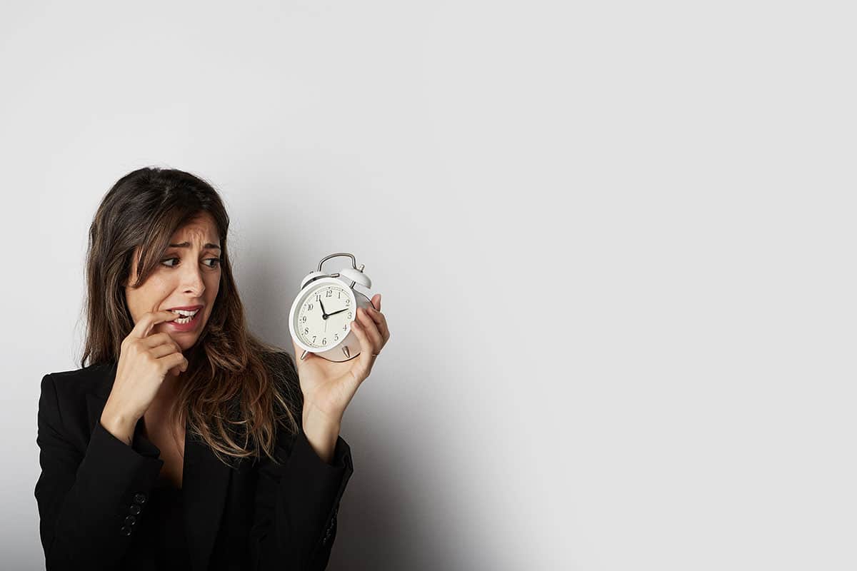 brunette woman dressed in black bites her finger while holding an old analog alarm clock