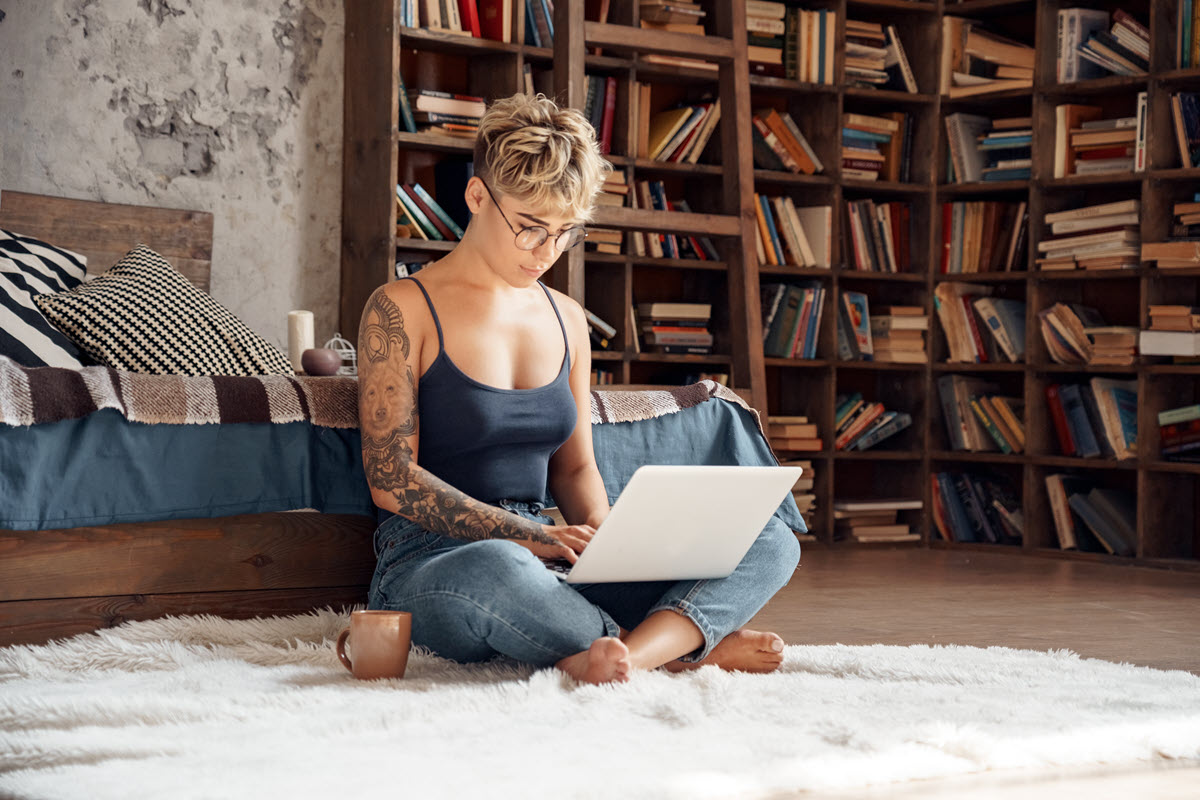 tattooed girl working on her laptop sitting cross legged on a carpet