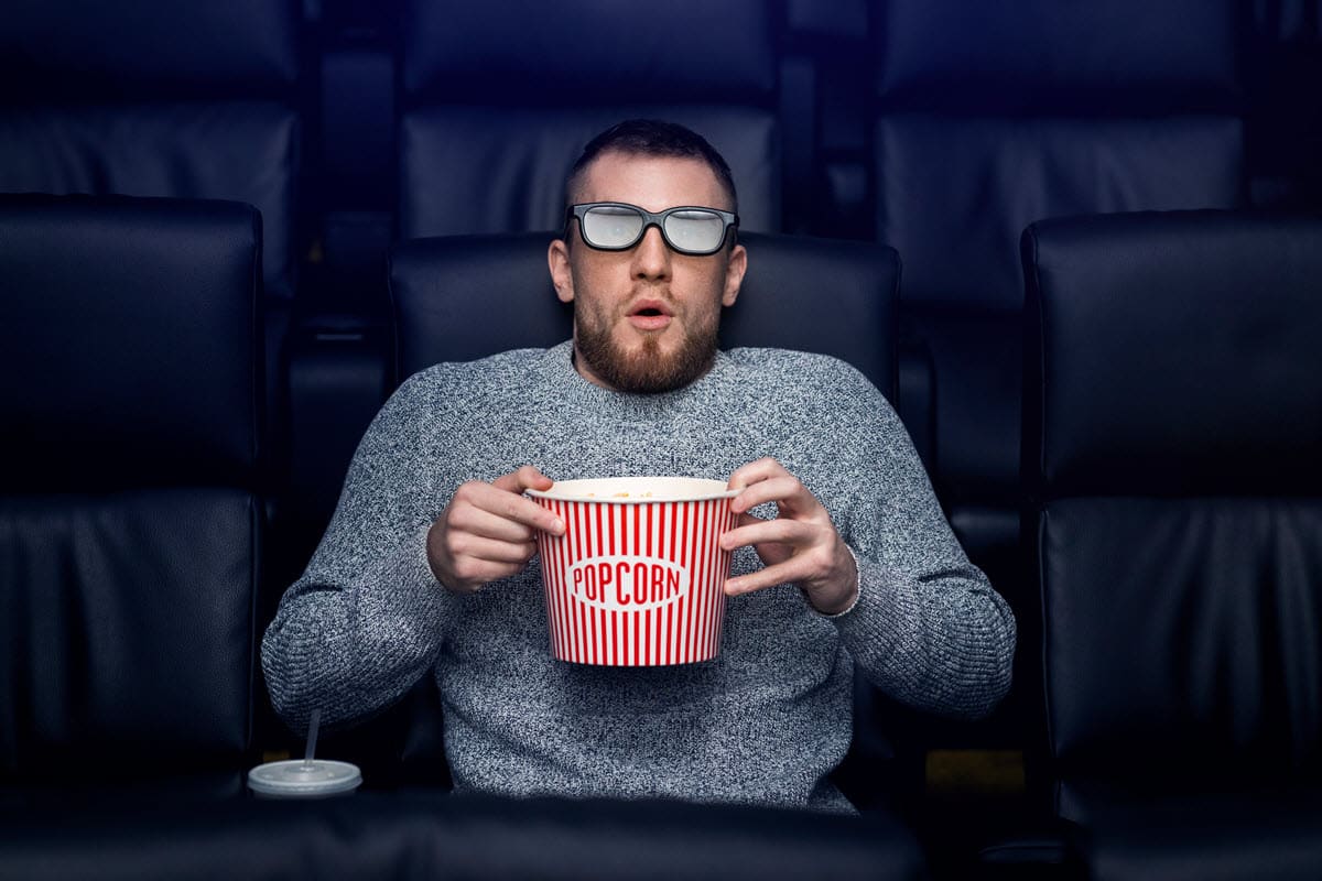 a man in a movie theatre sitting alone eating popcorn with 3d glasses on