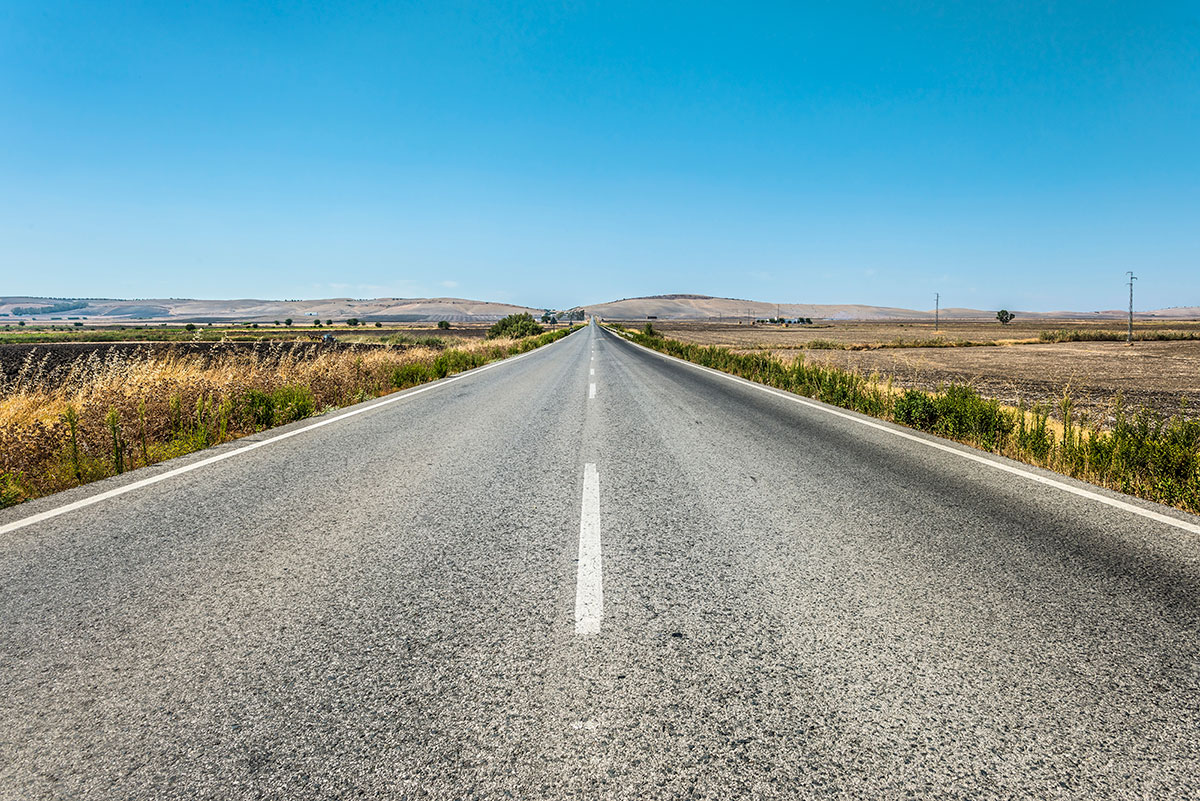 photo of a long road in summertime with lots of blue sky and rolling hills in the background