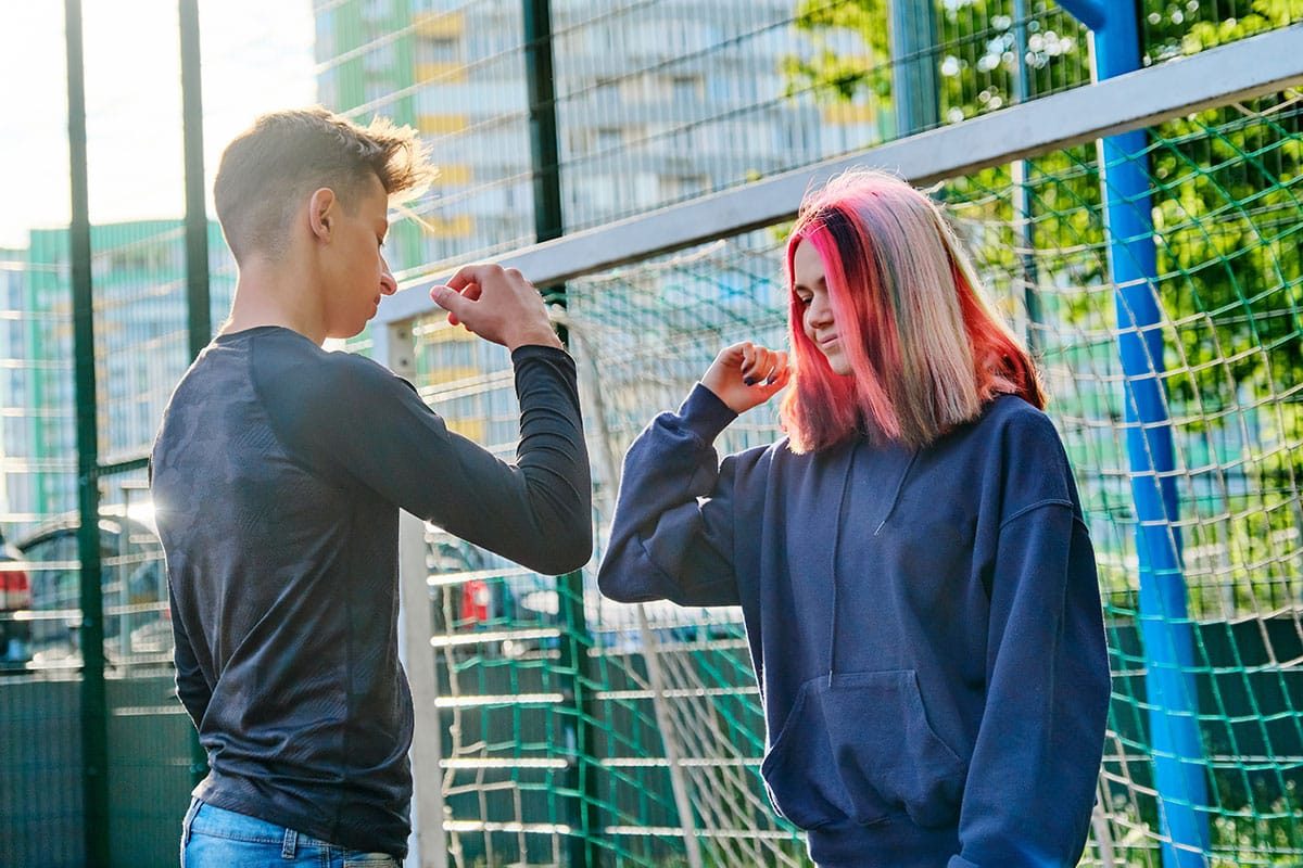 teenage boy and girl with pink dyed hair are bumping elbows in front of a soccer net as an excuse not to kiss someone