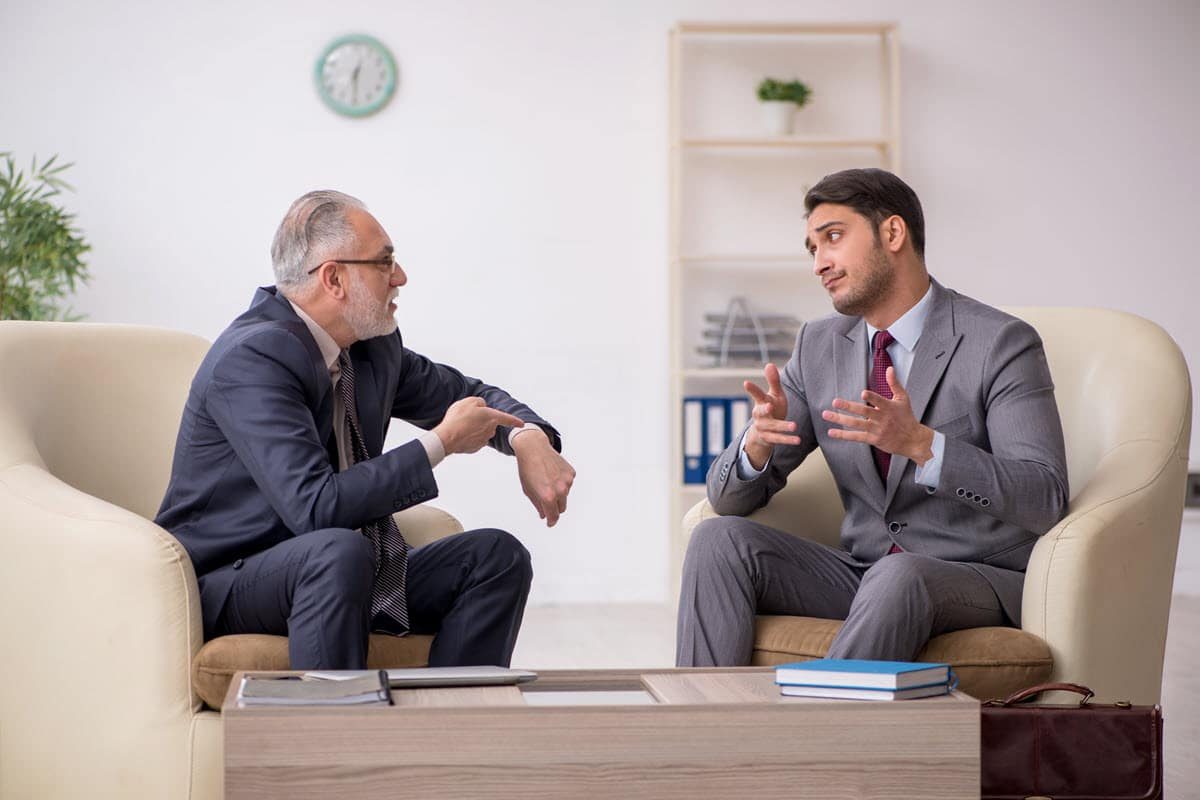 An older man pointing to his wrist indicating that the man he's talking to is late. Both men are wearing suits, sitting down in a professional setting.