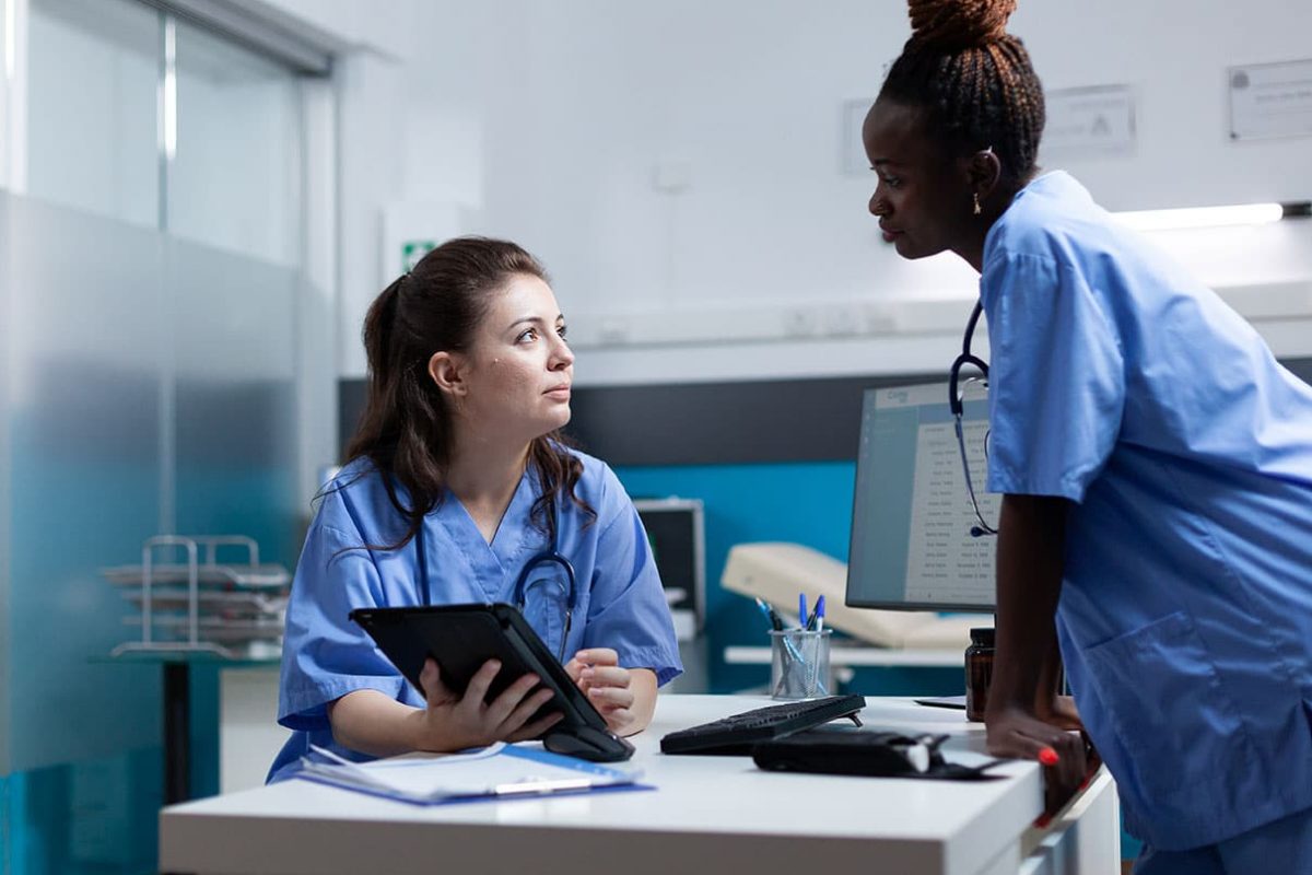 charge nurse standing over a desk at a clinic listening to another nurse give an excuse for nurses to call in sick