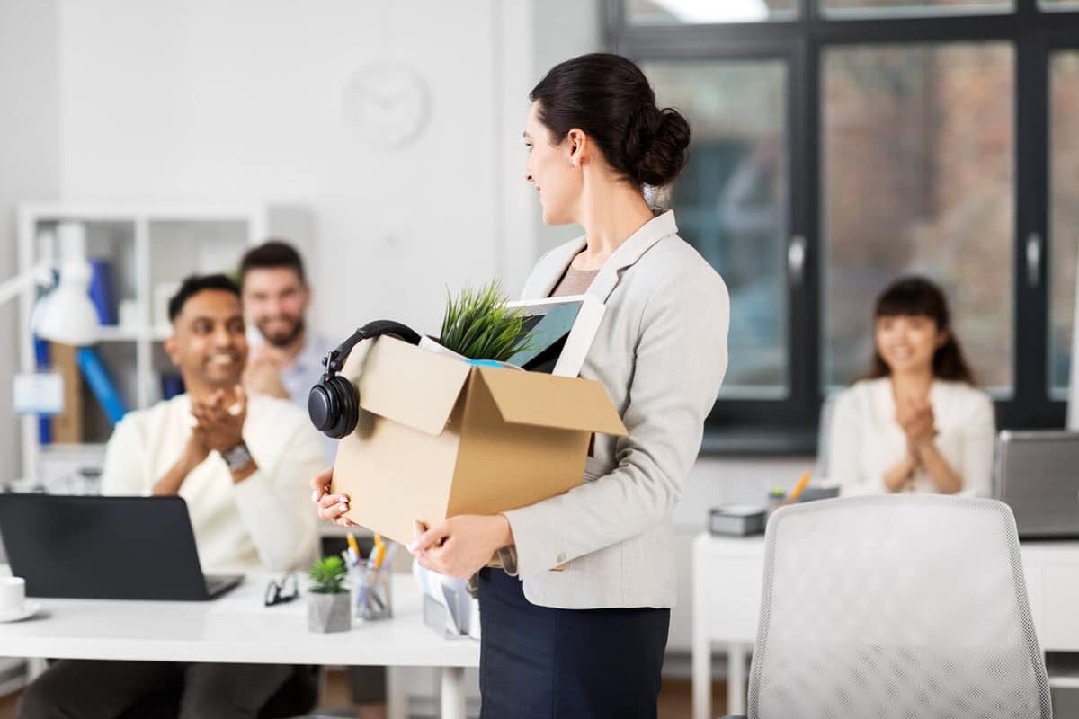 Woman who just quit her job is walking out of the building with a box and her belongings while her co-workers clap